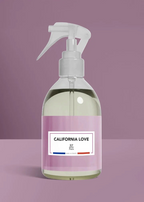 A clear plastic spray bottle with a white trigger sprayer and a pink label reading 'CALIFORNIA LOVE' and 'BP Paris', set against a solid mauve background.