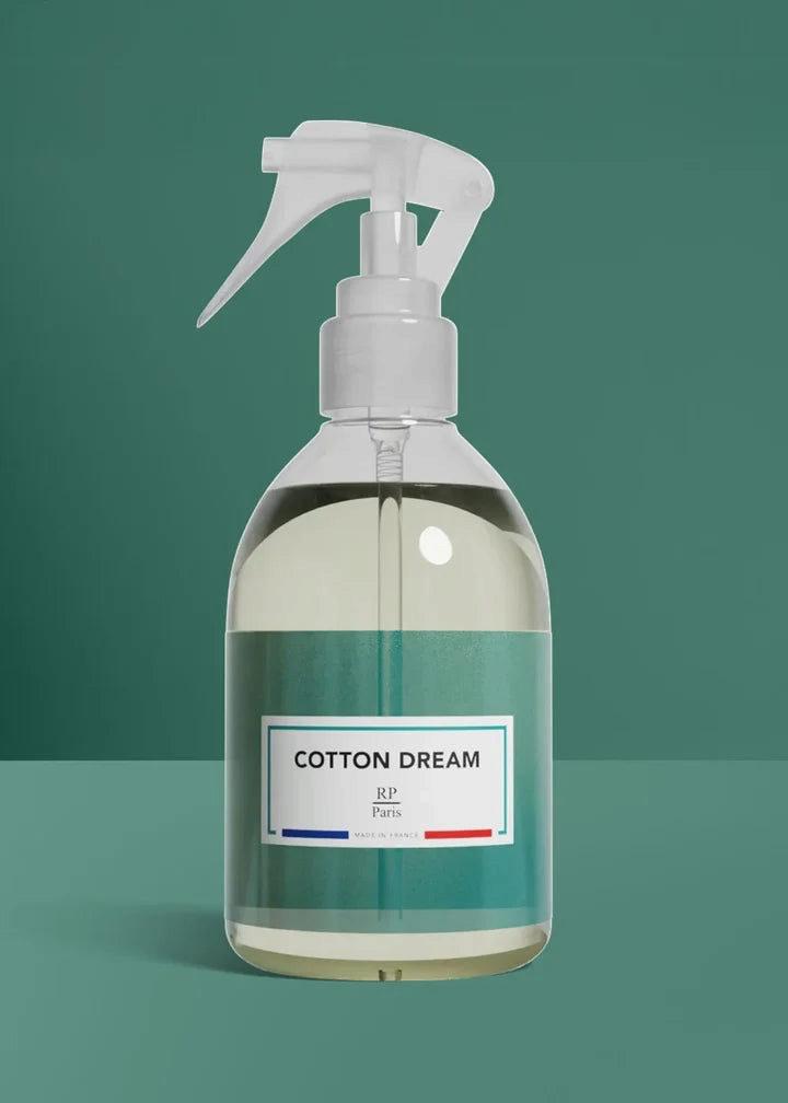 A clear plastic spray bottle filled with a pale yellow liquid, featuring a white trigger sprayer and a teal label with the text 'COTTON DREAM' and 'RP Paris' on a matching teal background.