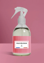 A clear plastic spray bottle filled with a pale yellowish liquid, featuring a white trigger sprayer and a pink label with the text 'FIGUE DÉLICIEUSE' and 'RP Farm'. The bottle is centered against a solid pink background.