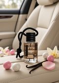 A small bottle of bubble gum fragrance with a black cord loop sits on a beige car seat, surrounded by pink and white gum balls, vanilla beans, and flower petals.