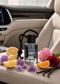A clear glass car diffuser bottle with a black cap and black hanging cord sits on a beige car seat, surrounded by orange slices, purple flowers, pink blossoms, a red rose, and vanilla pods. The background shows a modern car interior.