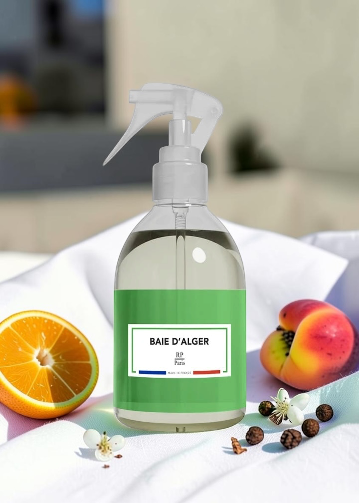A clear spray bottle with a green label reading 'BAIE D'ALGER' and 'Riviere' on the front, styled on white fabric with an orange slice, peach, and small flowers around it.