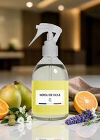 A clear spray bottle with a white trigger sprayer and a pale yellow label reading 'NÉROLI DE SICILE' and 'FR: Sicily', set on a countertop with fruit, lavender, and a towel in a blurred indoor kitchen-like environment.