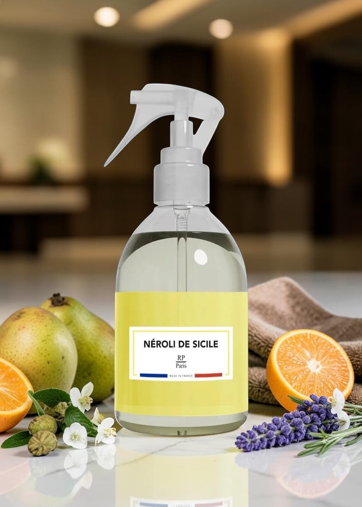 A clear spray bottle with a white trigger sprayer and a pale yellow label reading 'NÉROLI DE SICILE' and 'FR: Sicily', set on a countertop with fruit, lavender, and a towel in a blurred indoor kitchen-like environment.