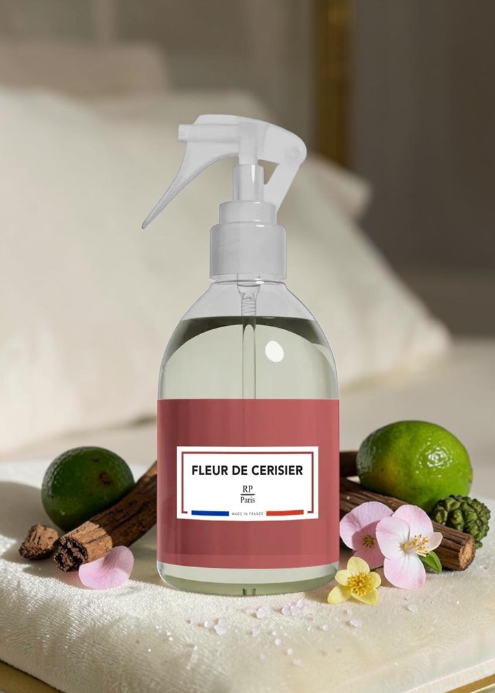 A clear spray bottle with a white trigger top and a dusty rose label reading 'FLEUR DE CERISIER' and 'BF Paris', placed on a towel with limes, cinnamon sticks, and pink petals around it.