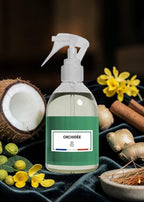 A clear spray bottle with a white trigger top and a green label reading ORCHIDÉE, surrounded by botanical props including a halved coconut, yellow flowers, cinnamon sticks, ginger root, and a bowl of incense on a dark velvet surface.