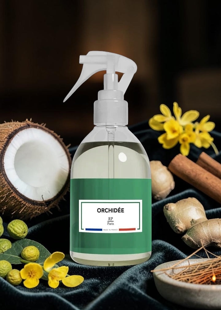 A clear spray bottle with a white trigger top and a green label reading ORCHIDÉE, surrounded by botanical props including a halved coconut, yellow flowers, cinnamon sticks, ginger root, and a bowl of incense on a dark velvet surface.