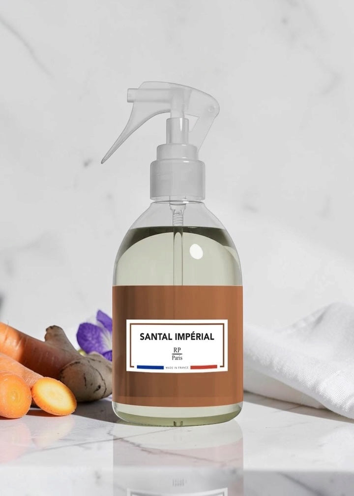 A clear plastic spray bottle filled with pale liquid, featuring a brown label with the text 'SANTAL IMPÉRIAL' and a small French flag graphic. The bottle is set on a white marble surface next to carrots, ginger, and a purple flower, with a white towel in the background.