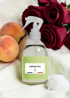 A clear spray bottle labeled 'ARABIAN TEA' with a pale green label and white trigger sprayer, styled on a white fabric surface with dark red roses, peaches, and a small pile of white salt crystals.