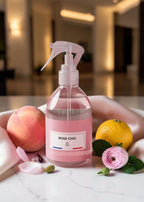 A clear spray bottle filled with pink liquid, labeled ROSE CHIC, styled on a marble surface with a peach, a lemon, a pink rose, and petals in a softly blurred indoor setting.