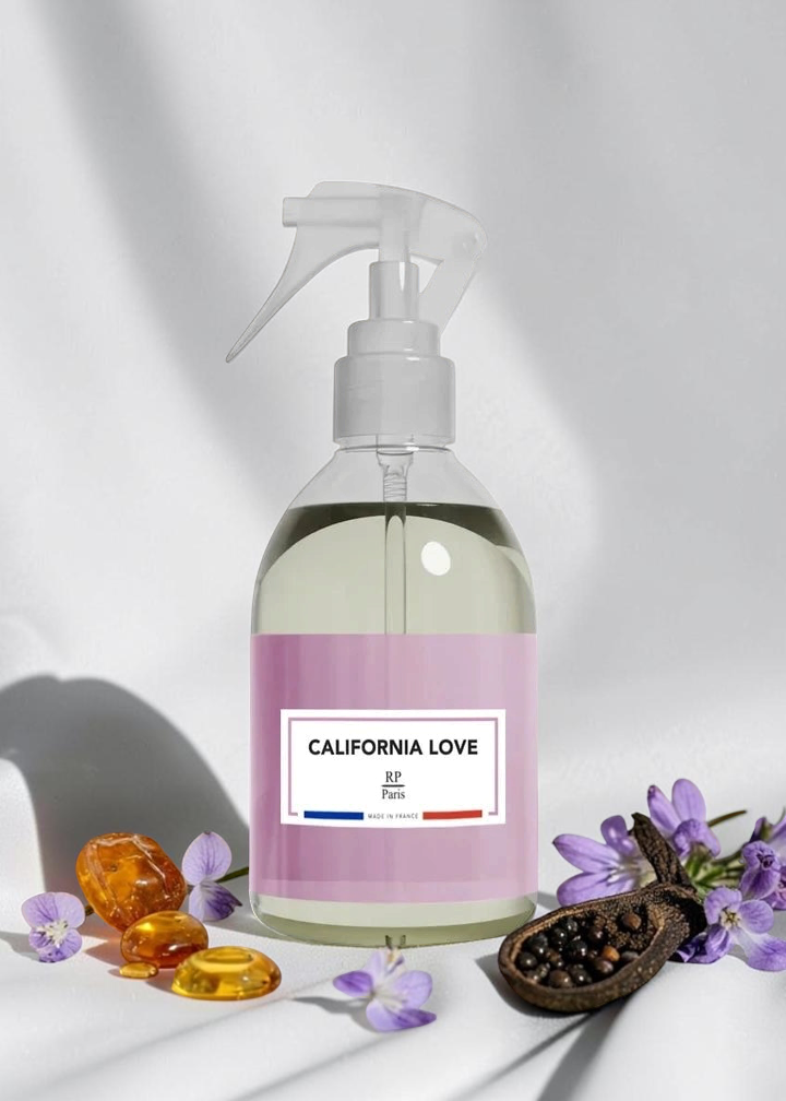 A clear spray bottle with a white trigger top and a lavender label reading 'CALIFORNIA LOVE' and 'by Pura', styled on a white surface with purple flowers, amber drops, and a pomegranate half around it.