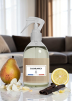 A clear plastic spray bottle with a white trigger sprayer and a beige label reading 'CASABLANCA' and 'Pérle', placed on a tabletop in a living room setting. A pear, lemon slice, vanilla pods, and flower petals surround the bottle.