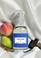 A clear pump bottle with a blue label reading 'CŌUR DE MUSC' and 'BF Paris' on a white satin background, styled with a peach, pear, cinnamon sticks, vanilla bean, and salt crystals.