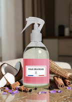 A clear spray bottle with a white trigger top and a pink label reading “FIGUE DÉLICIEUSE” and “Fragrance Oil,” styled on a countertop with coconut, cinnamon sticks, dried grass, and purple flower petals in a warm indoor setting.