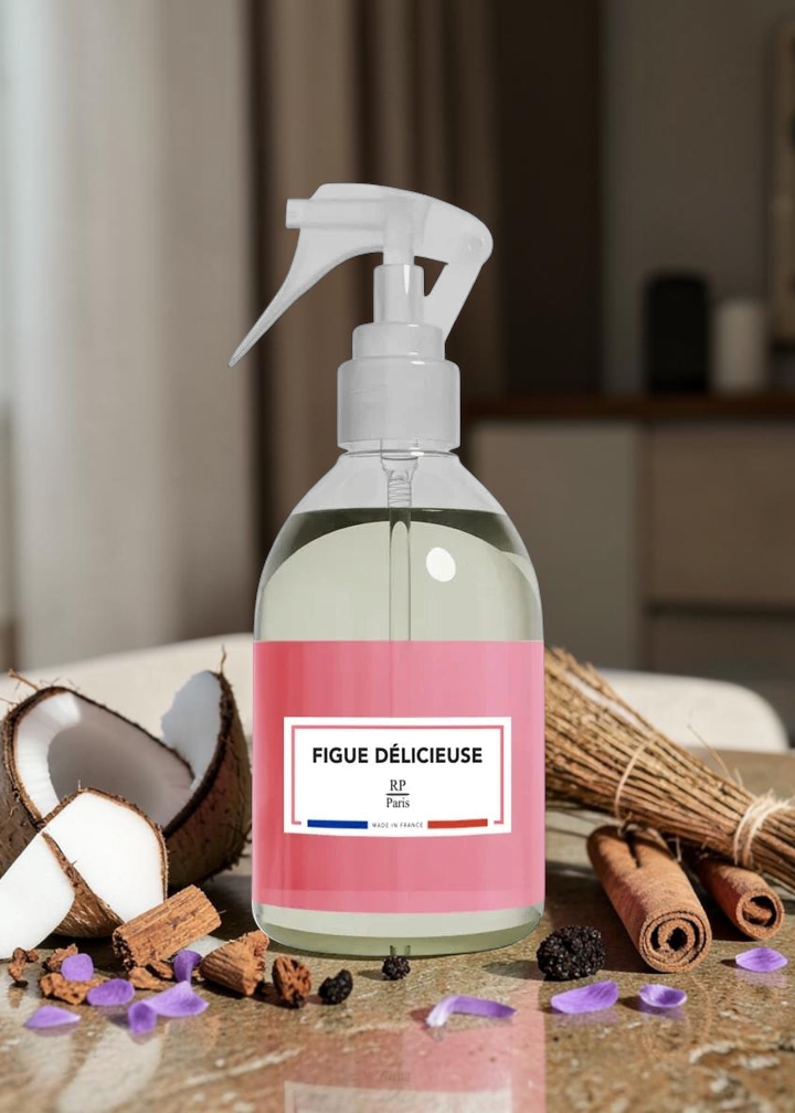 A clear spray bottle with a white trigger top and a pink label reading “FIGUE DÉLICIEUSE” and “Fragrance Oil,” styled on a countertop with coconut, cinnamon sticks, dried grass, and purple flower petals in a warm indoor setting.