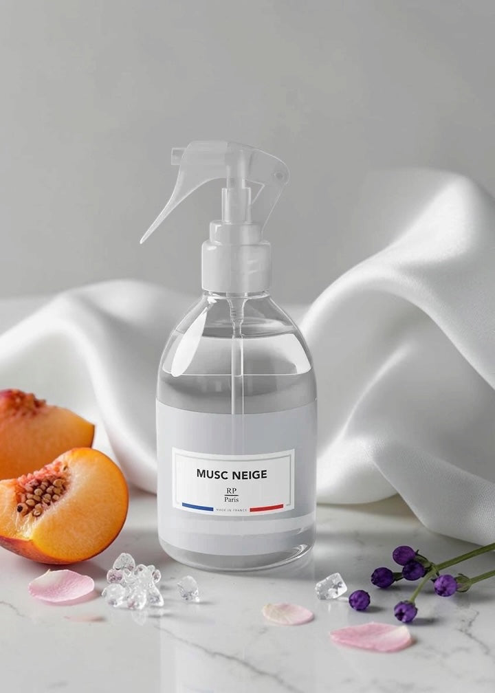 A clear spray bottle with a white trigger sprayer and a white label reading 'MUSC NEIGE' sits on a light marble surface. The bottle is styled with peach halves, pink petals, ice cubes, and small purple flowers against a soft white draped fabric background.
