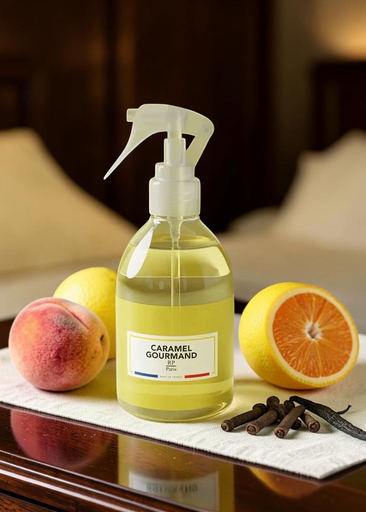 A clear spray bottle of Caramel Gourmand perfume oil with a white trigger sprayer and a white label, set on a table with fresh fruit including a peach, lemon, orange, and cloves against a warm, blurred background.