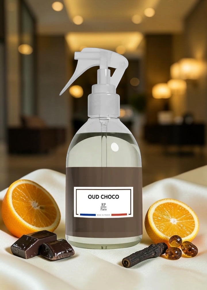 A clear spray bottle with a white trigger top and a brown label reading 'OUD CHOCO Paris' is centered on a draped white fabric surface. The bottle is surrounded by scent-related props: orange halves, dark chocolate pieces, and a cinnamon stick, with a softly blurred indoor background.