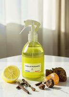 A clear spray bottle filled with pale yellow liquid stands on a white tabletop, labeled 'VANILLE ÉLÉGANTE' with a small French flag. A sliced lemon, cloves, orange segments, and brown sugar cubes are arranged around it in soft natural light.