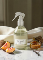 A clear spray bottle labeled 'COCO VANILLE' with a white trigger sprayer, filled with pale yellow liquid, styled on a marble surface with apricot slices, coconuts, and a small jar of amber liquid in the background.