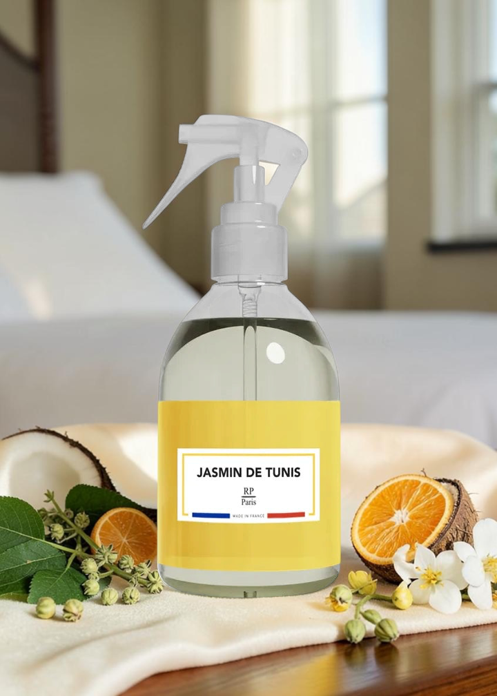 A clear spray bottle filled with pale liquid, featuring a white trigger sprayer and a yellow label that reads 'JASMIN DE TUNIS' with 'BF Paris' below. The bottle is styled on a cream towel with citrus slices, white flowers, and green leaves in a softly lit indoor setting.