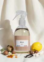 A clear pump bottle with a beige label reading BELLA and JP Perfum, filled with pale yellow liquid, styled on a draped cream fabric with a lemon, acorns, vanilla, and small white flowers around it.
