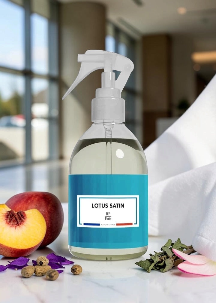 A clear spray bottle with a white trigger top and a blue label reading 'LOTUS SATIN Paris' sits on a marble surface. The bottle is styled with a peach, dried botanicals, and petals, with a blurred indoor background and a white towel.