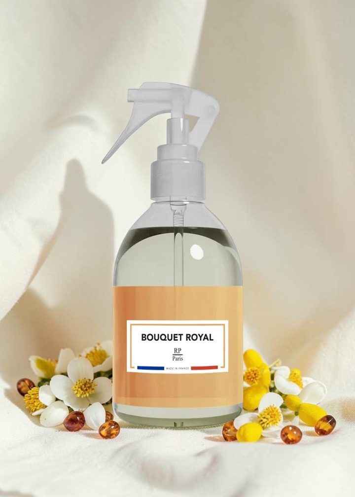 A clear spray bottle with a white pump dispenser and a peach-colored label reading 'BOUQUET ROYAL' and 'B5+ Perf', styled on draped cream fabric with white flowers and amber beads around its base.
