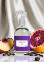 A clear spray bottle with a white pump and a purple label reading 'MUSC BLANC' and 'BF Paris', styled on a white surface with fruit slices, cloves, and cinnamon sticks against a draped light fabric background.