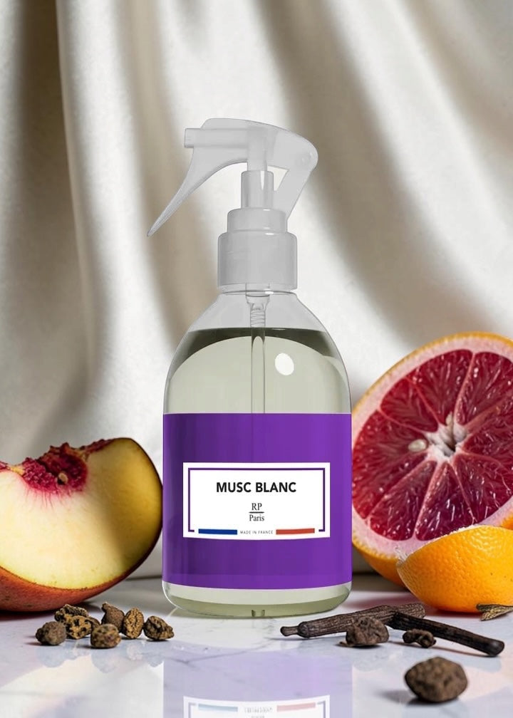 A clear spray bottle with a white pump and a purple label reading 'MUSC BLANC' and 'BF Paris', styled on a white surface with fruit slices, cloves, and cinnamon sticks against a draped light fabric background.