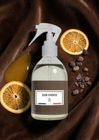 A clear spray bottle with a white trigger sprayer and a brown label reading 'OUD CHOCO' and 'BF Paris', styled on dark brown fabric with dried orange slices, amber liquid, and chocolate pieces.