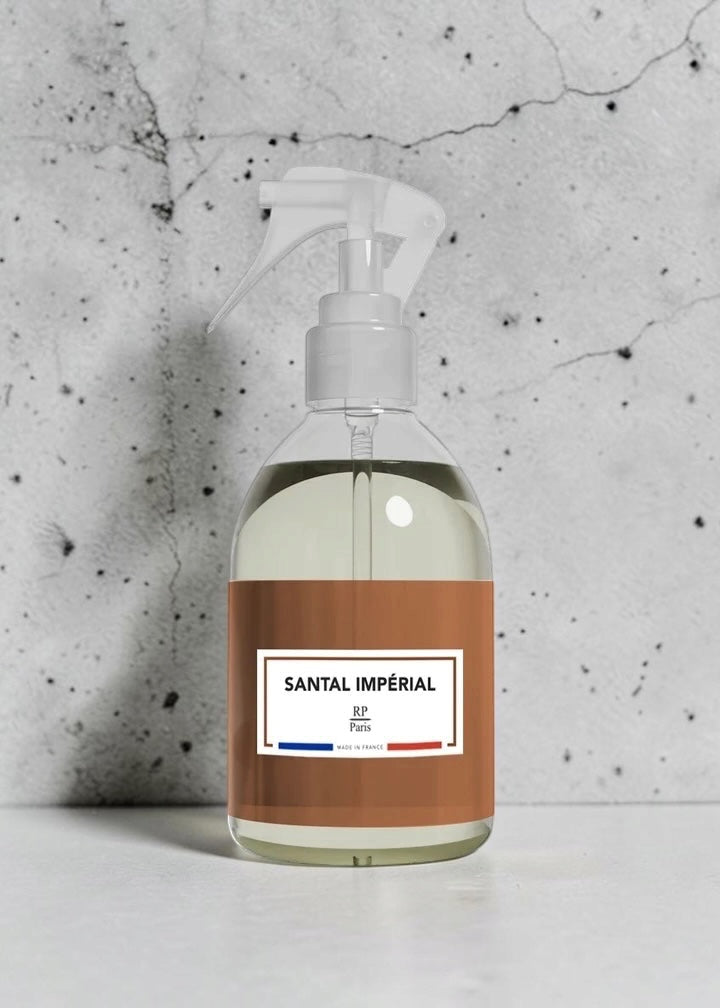 A clear plastic spray bottle filled with pale yellow liquid, featuring a white trigger sprayer and a brown label with the text 'SANTAL IMPÉRIAL' and a small blue and red accent, set against a textured gray concrete wall.