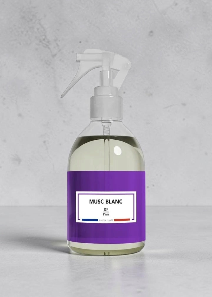 A clear plastic spray bottle filled with pale yellow liquid, featuring a white trigger sprayer and a prominent purple label with a white center panel that reads 'MUSC BLANC'.