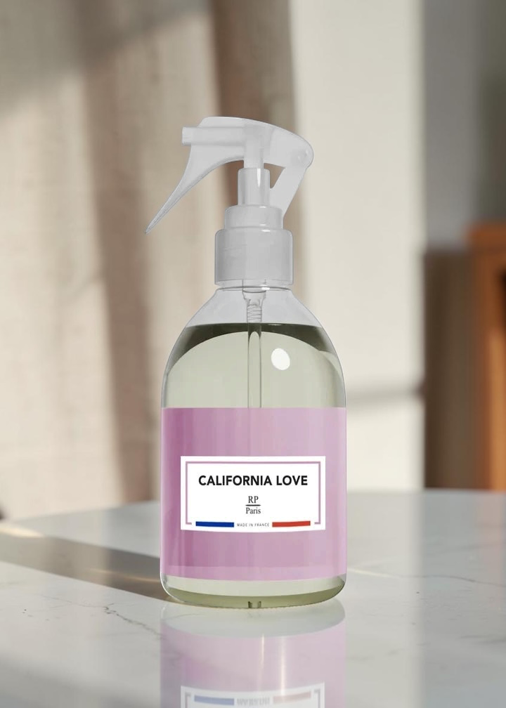 A clear trigger spray bottle filled with pale liquid, featuring a lavender label that reads 'CALIFORNIA LOVE' and 'SF, California.' The bottle is photographed on a glossy white surface with a softly blurred indoor background.