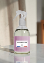 A clear trigger spray bottle filled with pale liquid, featuring a lavender label that reads 'CALIFORNIA LOVE' and 'SF, California.' The bottle is photographed on a glossy white surface with a softly blurred indoor background.