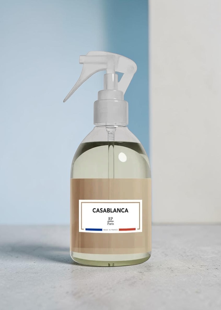 A clear plastic spray bottle filled with a pale yellow liquid, featuring a beige label with the text 'CASABLANCA' and a white trigger sprayer, set against a minimalist blue and white background.