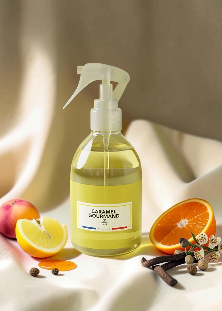 A clear plastic spray bottle filled with pale yellow liquid, labeled 'CARAMEL COUTMAND' with a small blue and red accent on the label, set on draped cream fabric with fruit and spice props including a lemon half, orange half, cinnamon sticks, and cloves.