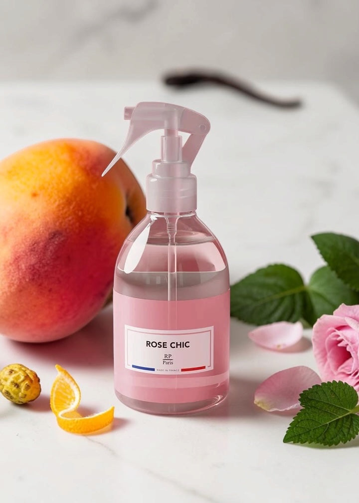 A clear spray bottle filled with pink liquid and labeled ROSE CHIC, styled on a light marble surface with a peach, pink rose, green leaves, and orange slices around it.