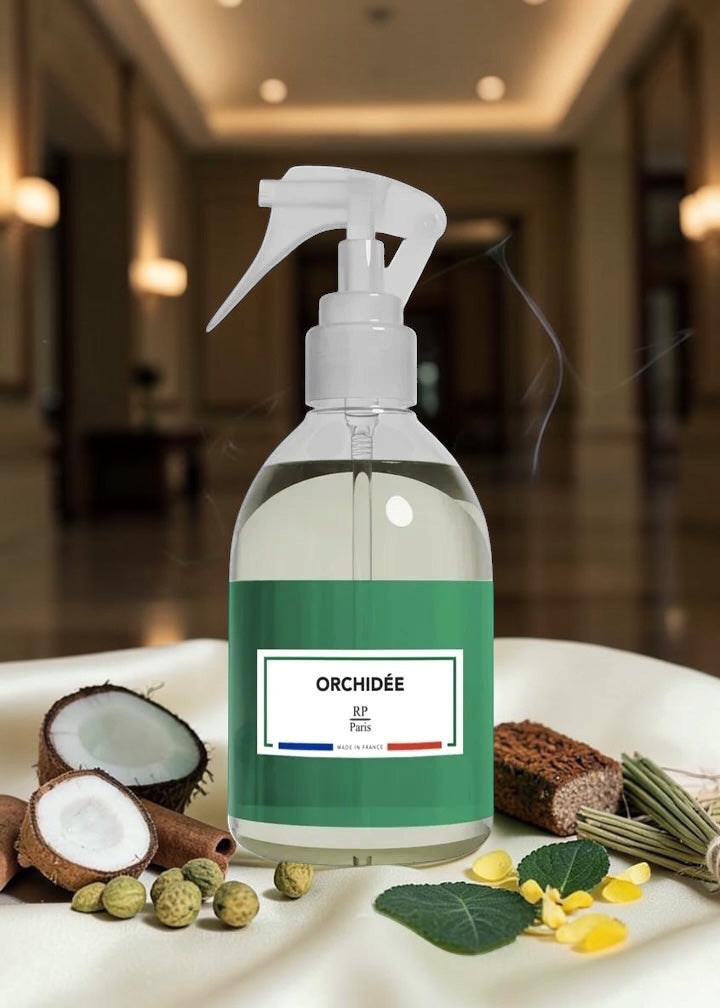 A clear spray bottle with a white trigger sprayer and a green label reading 'ORHIDÉE' and 'Paris', placed on a white surface with orchid-themed props including a coconut, green leaves, yellow petals, and dried flower stems. The background is a softly blurred indoor hallway.