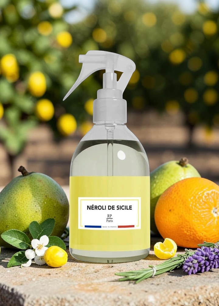 A clear spray bottle labeled 'NÉROLI DE SICILE' with a pale yellow liquid inside, styled on a stone surface with lemons, oranges, lavender, and rosemary, with a blurred citrus grove background.