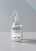 A clear glass spray bottle with a white trigger sprayer and a white label reading 'MUSC NEIGE' on a light gray surface against a pale blue-gray geometric background.