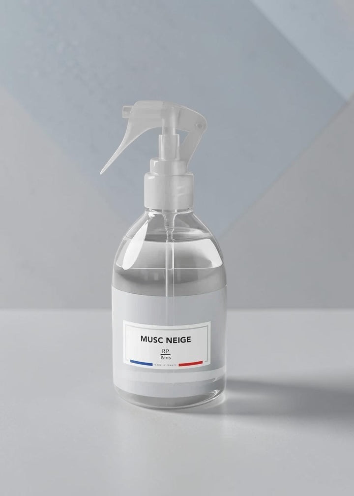 A clear glass spray bottle with a white trigger sprayer and a white label reading 'MUSC NEIGE' on a light gray surface against a pale blue-gray geometric background.